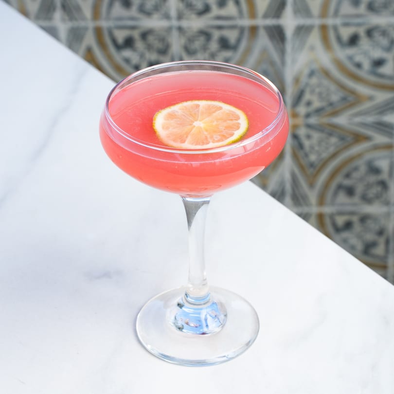 A vibrant pink cocktail with a lime slice garnish sits on a white marble table.