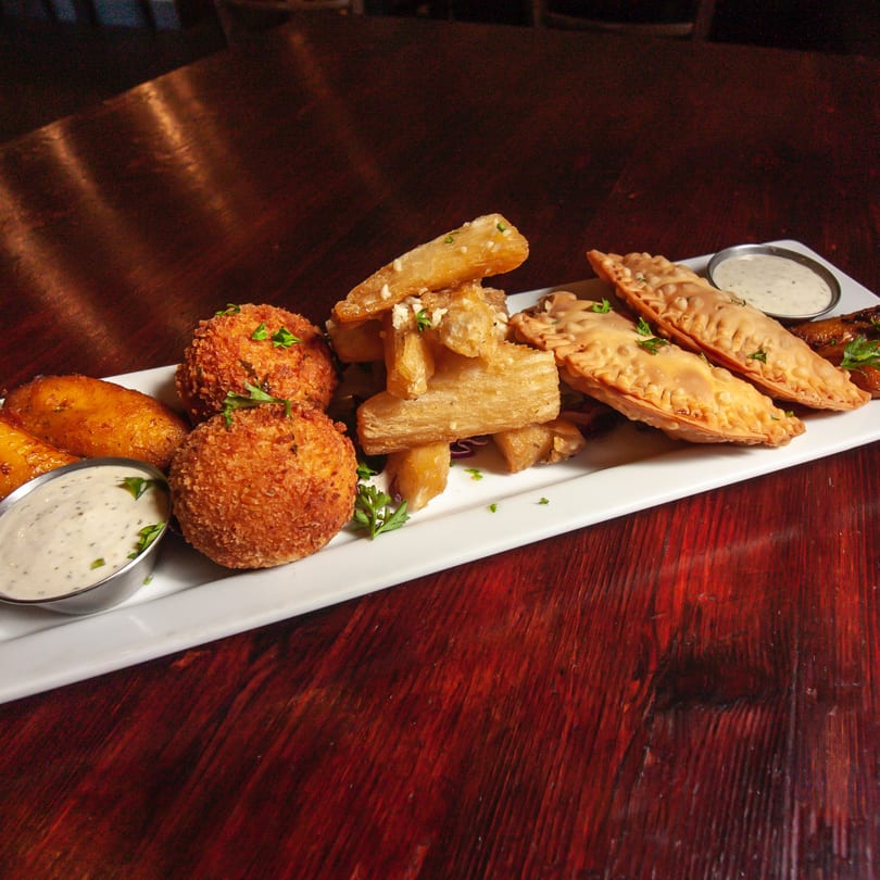 A white platter of fried plantains, croquettes, yuca fries, and empanadas with dipping sauces on a wooden table.