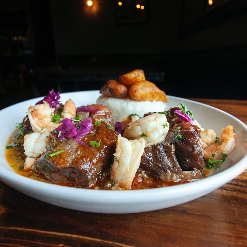 Braised short ribs and shrimp with rice, fried plantains, and purple cabbage on a white plate.