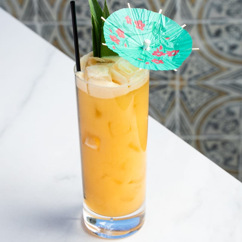Tall glass of orange cocktail with ice, a black straw, pineapple leaves, and a paper umbrella.