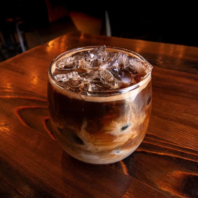 Iced coffee with milk swirls and ice cubes in a clear glass on a wooden table.