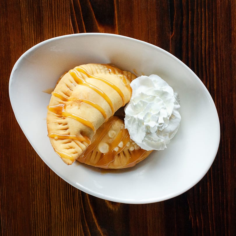 Two golden-brown empanadas drizzled with caramel sauce and a dollop of whipped cream in a white bowl.