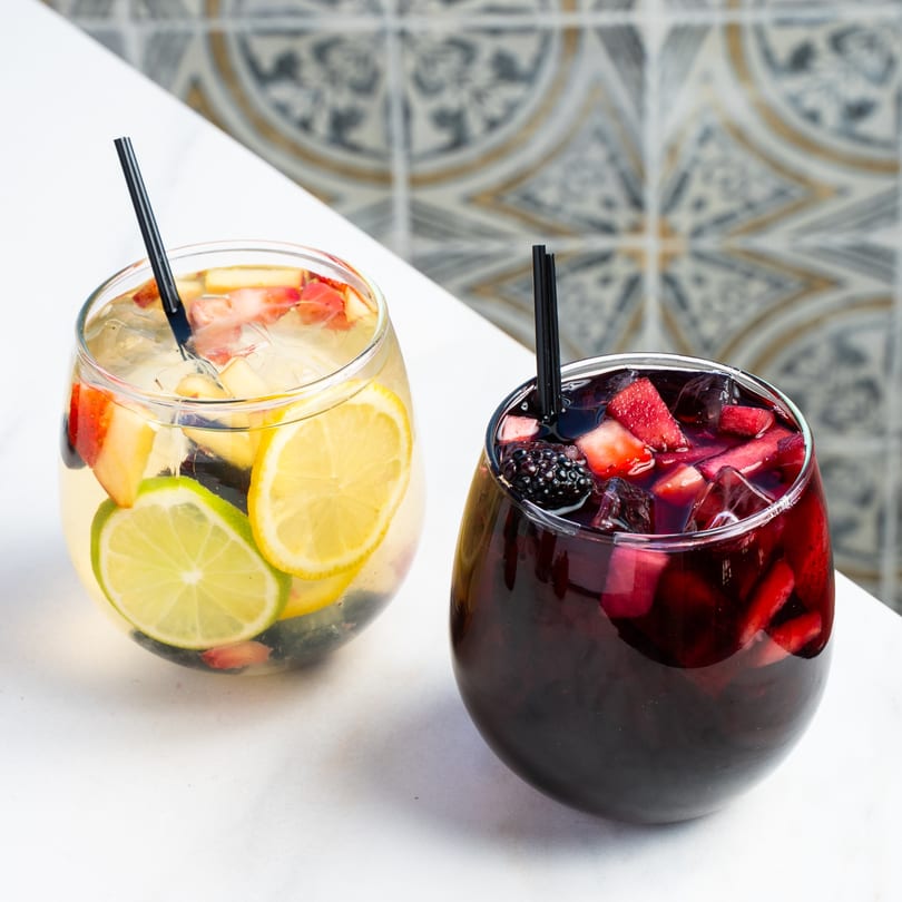 Two glasses of sangria, one white with citrus, one red with berries, on a white table.