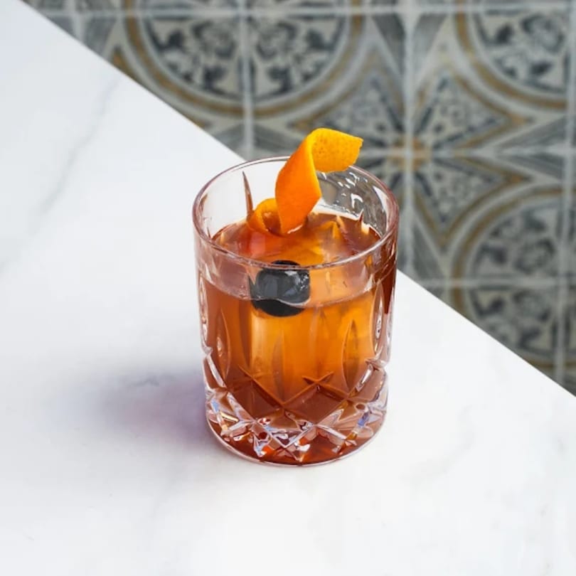 A Cuban cocktail in a cut glass with an orange peel and cherry on a white marble table.
